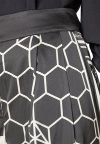 Jupe noire à finition brillante, avec un motif hexagonal blanc et des plis. La ceinture est lisse et satinée, ajoutant un contraste de texture.