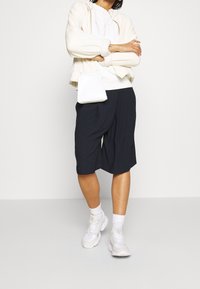 Chaqueta de forro polar blanca con cremallera, sudadera blanca con gráfico, pantalones cortos azul marino hasta la rodilla, combinados con zapatillas blancas y un pequeño bolso bandolera.