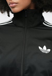 Mustast siledast kangast lukuga jope, millel on valged kolmetriibulised detailid õlgadel ning valge Adidas logo rinnal.