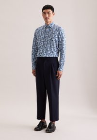 Camicia a maniche lunghe blu con motivo floreale bianco abbinata a pantaloni plissettati blu navy e scarpe di pelle nere. Tessuto strutturato.