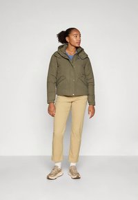 Alyvuogių žalios spalvos pūkinė striukė su gaubtu, priekyje susegama spaustukais ir dviem įstrižomis kišenėmis. Dėvima ant mėlynos spalvos marškinių, derinama su smėlio spalvos kelnėmis ir rudos spalvos sportbačiais.