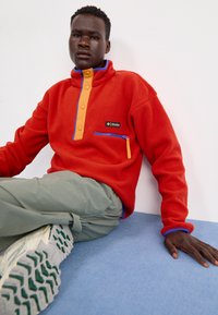 Rød fleecepullover med en blå og orange lynlåslomme, orange knaplukning og blå kraveakcenter. Grå bukser og cremefarvede sneakers.