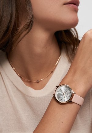 Frau trägt eine roségoldene Uhr mit einem pinkfarbenen Armband und einem Zifferblatt mit Baum-Design sowie eine zarte goldene Halskette mit kleinen Perlen.