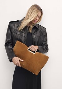 Borsa a busta in suede marrone con manico rettangolare dorato, tenuta da una persona in giacca di pelle nera e abito nero.