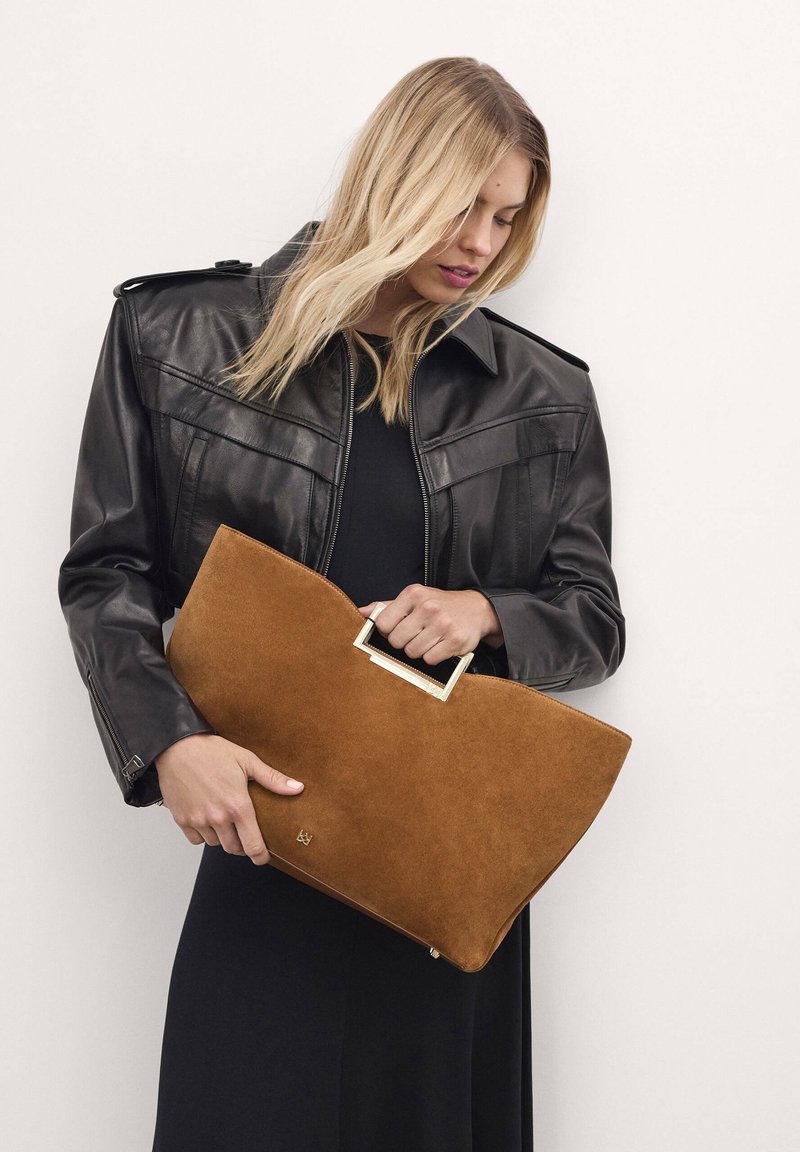 Borsa a busta in suede marrone con manico rettangolare dorato, tenuta da una persona in giacca di pelle nera e abito nero.