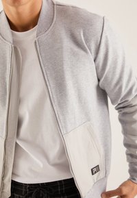 Chaqueta bomber de color gris claro, fabricada con una tela suave, que cuenta con un cierre de cremallera en la parte delantera, bolsillos contorneados y acentos en beige en contraste.