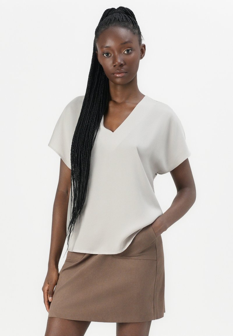 Blusa de cuello en V gris claro con mangas cortas, superpuesta sobre una mini falda marrón. Tela suave con un brillo sutil. La modelo posa con las manos en los bolsillos.