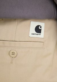 Bežne Carhartt hlače s teksturom tkanine, s zatvaranjem na gumb i bijelom oznakom s Carhartt logotipom na pojasu.