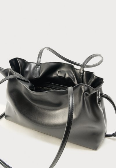 Μαύρη τσάντα tote από μαλακό δέρμα με λεπτούς ιμάντες ώμου και ανοιχτό πάνω μέρος που δείχνει μια μικρή εσωτερική τσέπη με φερμουάρ.