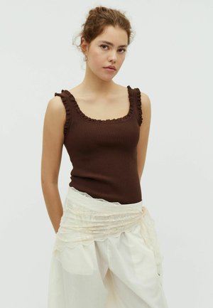 Young woman with curly hair wears a sleeveless brown ribbed top and white pants with a sheer lace wrap, standing against a plain background.