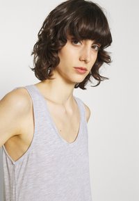 Šviesiai pilkas be rankovių marškinėlis su V-formos iškirpte. Minkšta, tekstūruota medžiaga su laisvu siluetu ir plačiomis išpjovomis rankovėms.