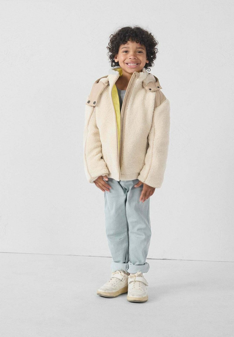 Chaqueta de forro polar en color crema con detalles en tono marrón claro, cierre de cremallera y botones en los hombros. Combinada con pantalones azul claro y zapatillas blancas.