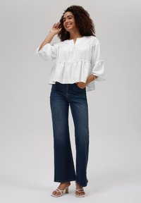 Hvit, teksturert bluse med v-hals og volangdetaljer, sammen med mørkeblå utsvingte jeans. Modell som har på seg hvite sandaler med stropper.