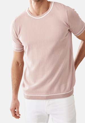 Mand iført en lyserød T-shirt med korte ærmer og hvide detaljer, parret med hvide bukser mod en ensfarvet baggrund.