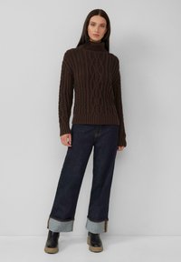 Dunkelbrauner Strickrollkragenpullover mit einem Zopfmuster, kombiniert mit weiten dunkelblauen Jeans aus Denim mit kontrastierenden hellblauen Säumen.