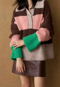 Femme portant un pull en laine coloré avec des manches marron, rose, beige et vert, associé à une mini-jupe en cuir marron foncé.