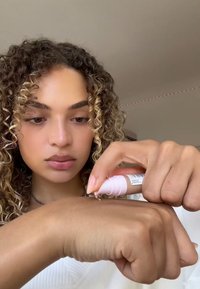 Un applicatore per siero rosa che distribuisce un prodotto cremoso su un avambraccio abbronzato. Lo sfondo presenta un'illuminazione soffusa e una tonalità neutra.