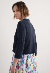 Temno moder skrajšan kardigan s 3/4 rokavi, okroglim izrezom in rebrastim robom. Nosi se prek obleke s cvetličnim vzorcem v živih barvah.