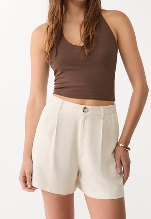Femme portant un short en lin beige taille haute avec un bouton et des passants de ceinture, associé à un top court marron sans manches et un bracelet en or.