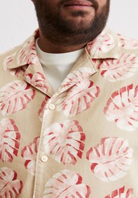 Korte mouwen shirt in beige stof met grote rode en roze bladpatronen, voorzien van een kraag en witte knopen. Binnenlaag is een eenvoudige witte t-shirt.