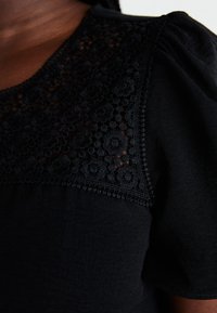 Top noir avec un panneau en dentelle florale texturée à bord festonné et des manches bouffantes, fabriqué dans un tissu léger.