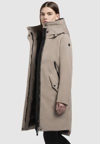 Beige lange jas met een hoge kraag, een rits aan de voorkant, zijzakken en zwarte accenten. Glad materiaal, ontworpen voor warmte.