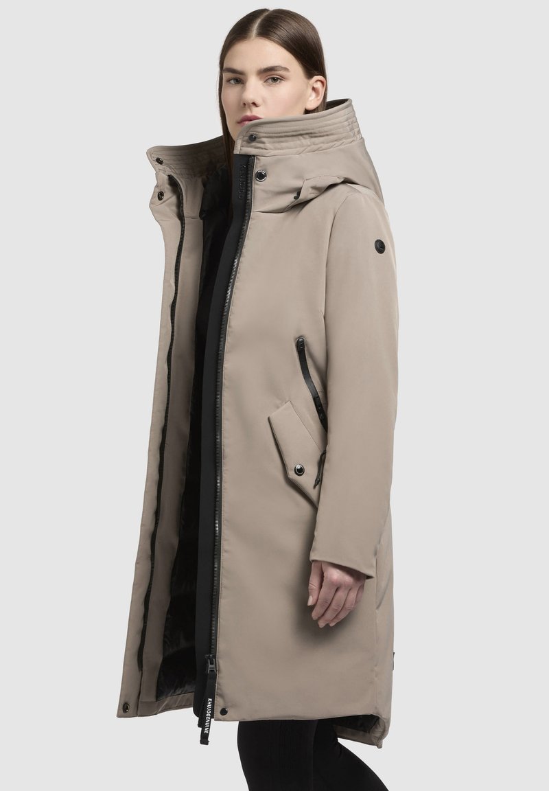 Cappotto lungo beige con colletto alto, chiusura frontale con zip, tasche laterali e dettagli neri. Texture liscia, progettato per il calore.