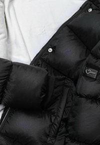 Chaqueta acolchada negra con un acabado brillante, puños acanalados y una etiqueta de marca prominente. Presenta un cierre de cremallera con broches a presión y secciones acolchadas.