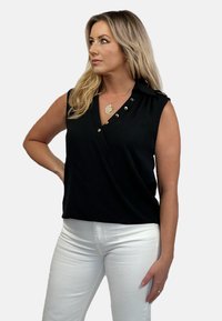 Czarny bezrękawnik z krojem w stylu wrap, z srebrnymi guzikami na ramieniu i wzdłuż dekoltu, zestawiony z białymi spodniami.