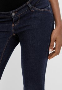 Jeans de mezclilla de un azul oscuro con costuras naranjas, diseño de cinco bolsillos y una cintura de altura media. La toma en primer plano muestra la textura y el ajuste alrededor del muslo.