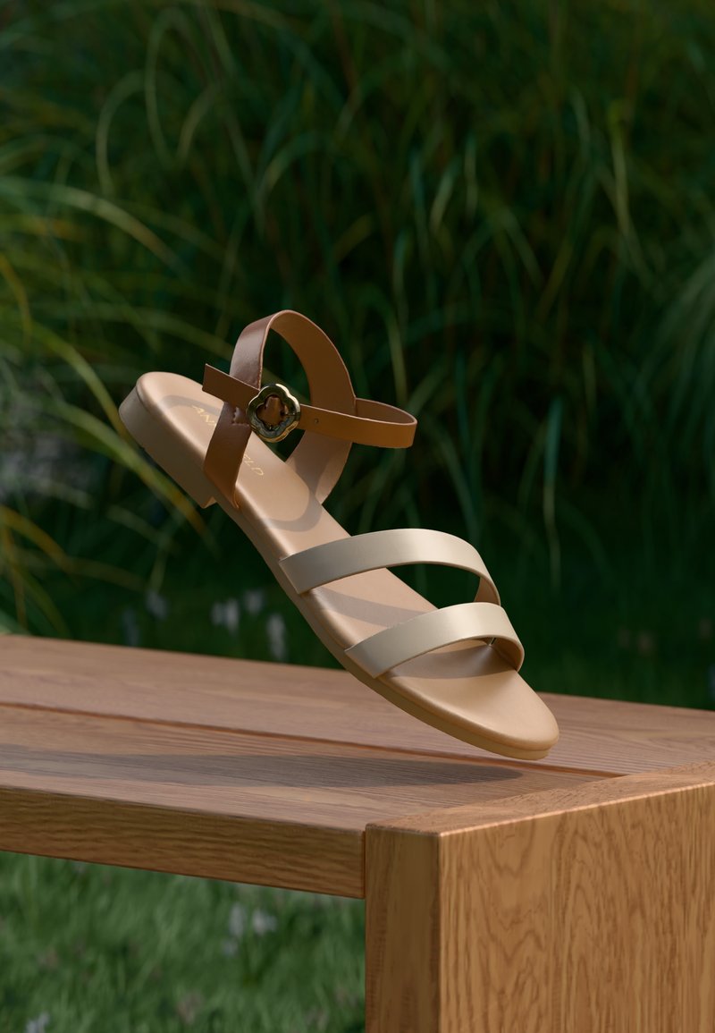 Μπεζ επίπεδη σανδάλι με τρεις πλατιές λουράκια και καφέ λουράκι στον αστράγαλο. Διακοσμημένο με αγκράφα σε χρυσή απόχρωση με floral σχέδιο. Λείο συνθετικό υλικό.