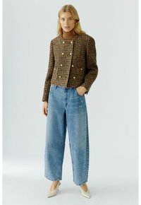 Blonde vrouw in een bruine geruite jas over een bruine coltrui, met wijde lichtblauwe jeans en witte puntige hakken, staand met één hand in haar zak.