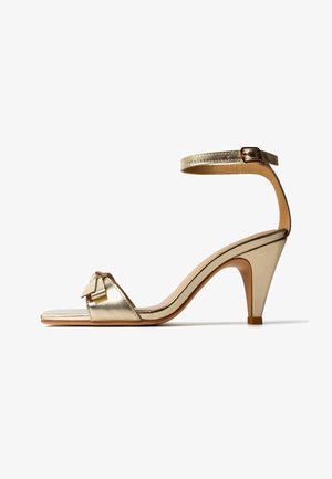 Gold high-heeled sandal with ankle strap, open toe, and decorative bow on front strap, shown on a white background.