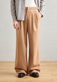 Pantalones anchos beige con pliegues frontales, un lazo para cinturón y bolsillos laterales, combinados con una camisa a cuadros azul y amarilla y zapatos negros.
