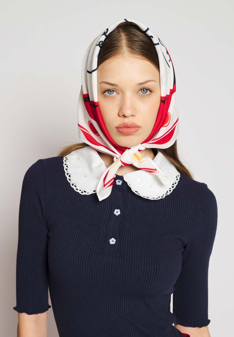 Jeune femme aux yeux bleus portant un foulard rouge et blanc à motifs et un haut bleu marine avec un col blanc festonné.