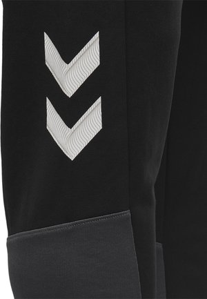 Czarne dresy z szarym elastycznym mankietem i dwoma ukośnymi wzorami chevron w teksturowanym jasnoszarym kolorze po bokach.