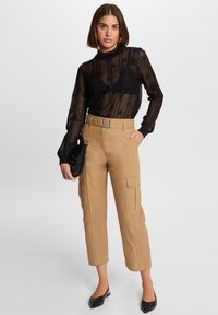 Zwarte doorzichtige top met lange mouwen en een textuurpatroon, gecombineerd met beige cargobroek met zijzakken en een brede riem. Zwarte handtas.