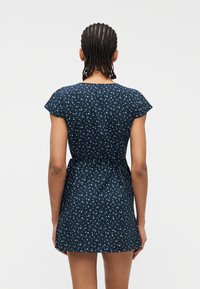 Άτομο με κοτσίδες που φαίνεται από πίσω, φοράει κοντομάνικο μπλε-σκούρο φόρεμα με μικρά λευκά λουλουδάτα σχέδια και ζώνη στη μέση.