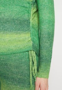 Sweater verde de malha com um gradiente do verde escuro para o verde claro, apresentando punhos canelados e laços laterais. Tecido suave e texturizado.