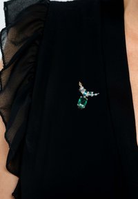 Broche de gema verde sobre una tela negra con bordes rizados. La broche presenta acentos azules y un diseño curvado decorativo.
