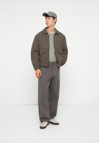 Brown checkered jacket, green crewneck, gray wide-leg pants, and white sneakers with brown accents. Model wears a matching cap.