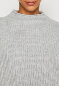Detailní záběr na osobu ve světle šedém žebrovaném pleteném svetru s vysokým výstřihem a měkkou texturou.