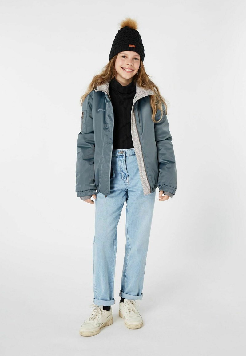 Leichte, graue Daunenjacke mit einem fleecegefütterten Innenfutter. Schwarze Strickmütze mit einem braunen Etikett. Blaue Jeans und weiße Sneaker vervollständigen das Outfit.