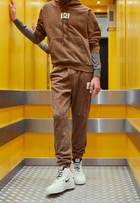 Sudadera marrón con un bolsillo frontal y un logo, combinada con joggers texturizados a juego y zapatillas blancas, ambientada en un ascensor amarillo.