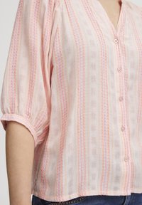 Blusa rosa chiaro con scollo a V, maniche a sbuffo corte e chiusura con bottoni. Presenta strisce verticali in tonalità arancioni e rosa con un motivo testurizzato.