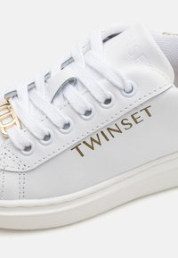 Sneaker in pelle bianca con logo dorato "TWINESET", lacci bianchi, occhielli rotondi e suola in gomma testurizzata. Design pulito con minimi dettagli.