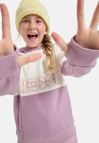 Sudadera morada y crema con un diseño gráfico, que presenta mangas sin dedos y un detalle de pelo trenzado. El modelo lleva un gorro de punto amarillo.