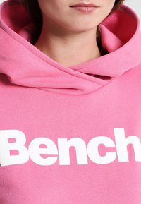 Sudadera rosa hecha de un tejido suave, con un gran logo blanco "Bench" en la parte frontal. La capucha tiene un diseño relajado y caído.