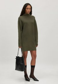 Olivgrünes Strickkleid mit hohem Kragen und oversized Passform, kombiniert mit schwarzen Lackleder-Pumps und einer schwarzen Tragetasche.