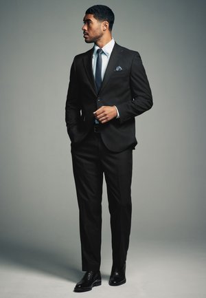 Hombre de pie, vistiendo un traje negro, corbata azul, camisa blanca y zapatos negros, mirando hacia su izquierda contra un fondo gris liso.